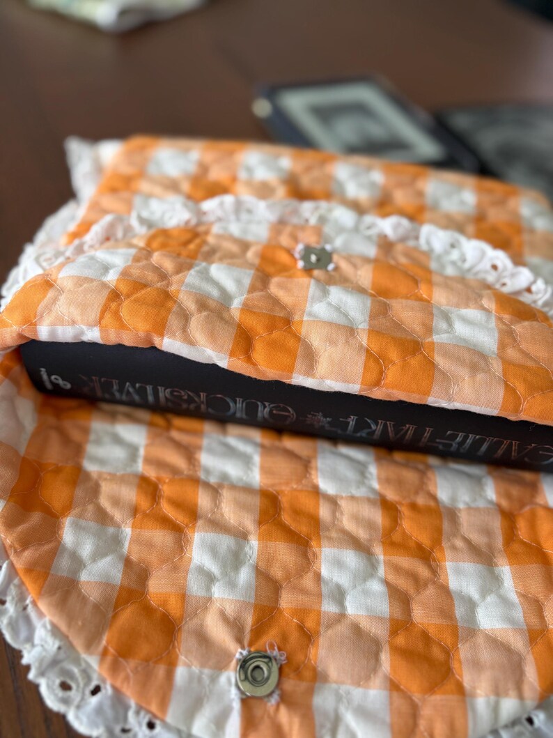 May include: An orange and white gingham quilted book sleeve with a white lace trim. The sleeve is closed with a silver snap. A book with the title "GALILEO'S TART QUICKSILVER" is inside the sleeve.