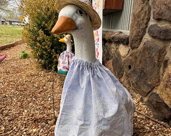 May include: A concrete goose statue wearing a straw hat with a floral band and a light blue dress with a white floral pattern. Another goose statue is visible in the background, also wearing a hat and dress.