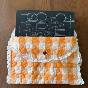 May include: A quilted book sleeve in orange and white gingham pattern, with a white lace trim. The sleeve holds a book with the title "Social Cues" visible. A small, dark red button secures the flap.