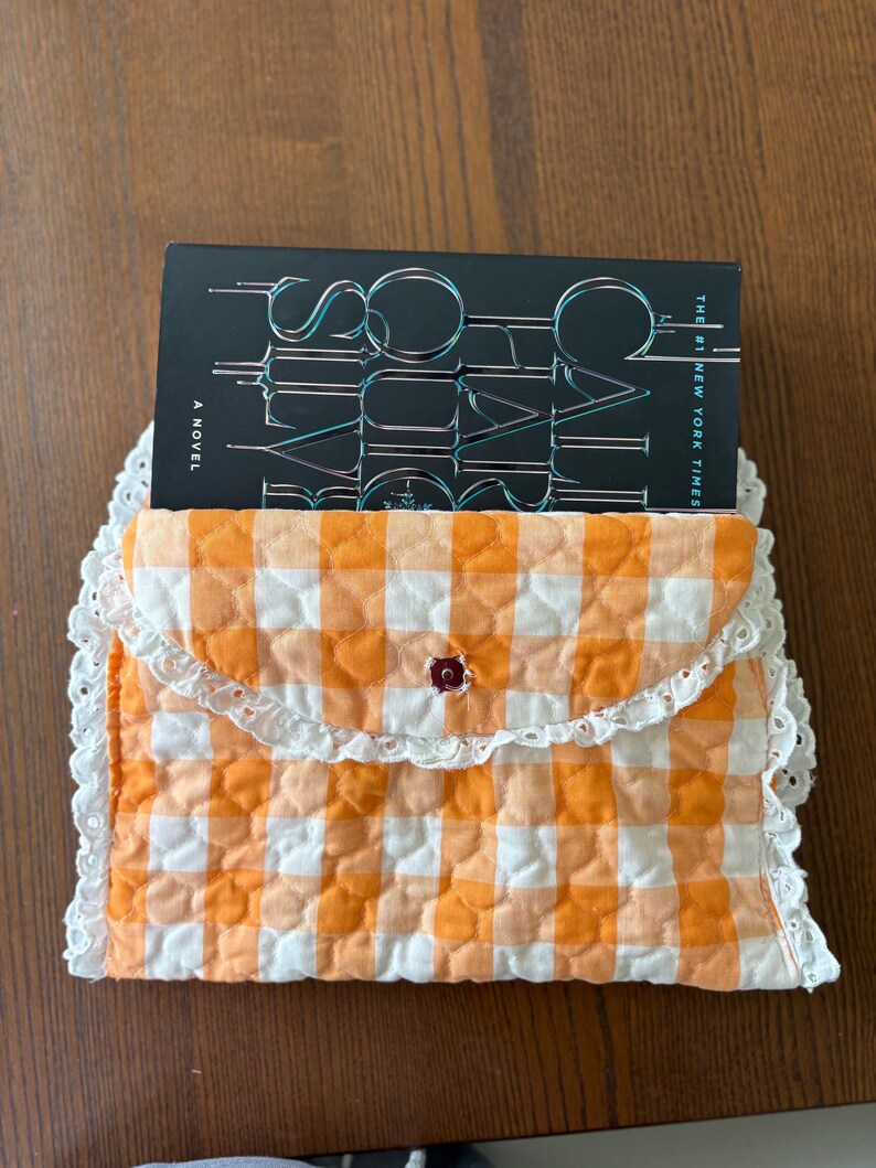 May include: A quilted book sleeve in orange and white gingham pattern, with a white lace trim. The sleeve holds a book with the title "Social Cues" visible. A small, dark red button secures the flap.