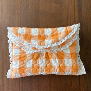 May include: A quilted clutch with an orange and white gingham pattern. The clutch is rectangular with a scalloped white lace trim. The fabric has a quilted texture, and the color scheme is reminiscent of a picnic blanket.