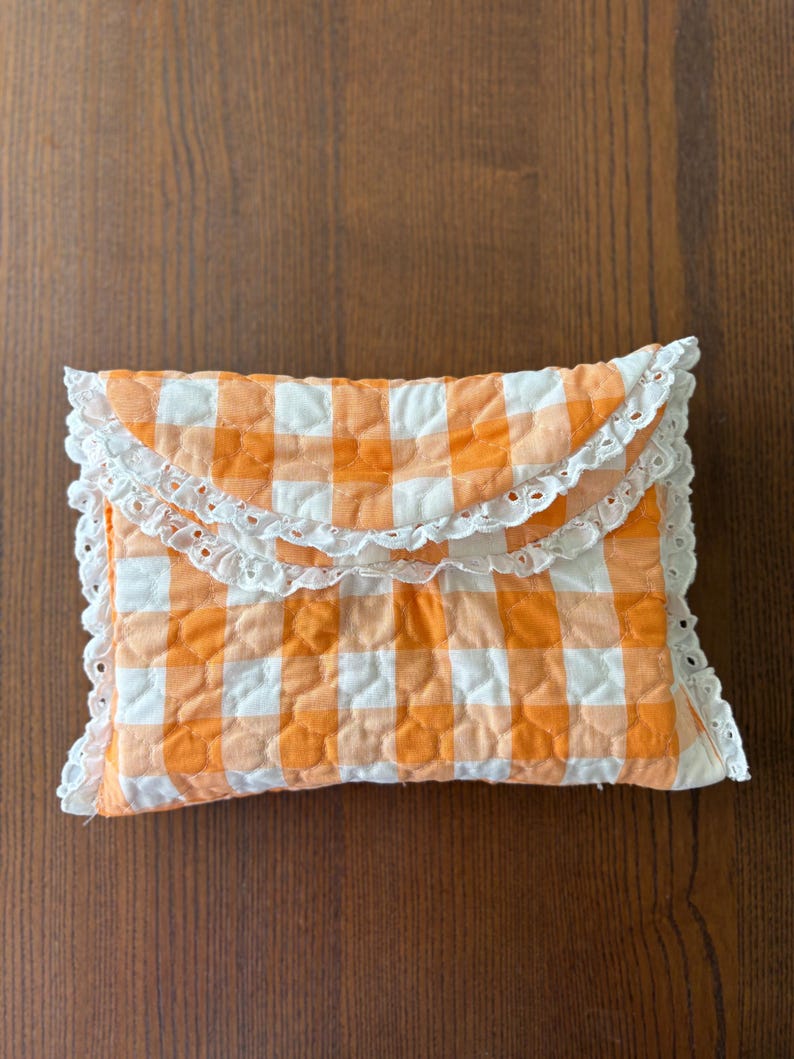 May include: A quilted clutch with an orange and white gingham pattern. The clutch is rectangular with a scalloped white lace trim. The fabric has a quilted texture, and the color scheme is reminiscent of a picnic blanket.