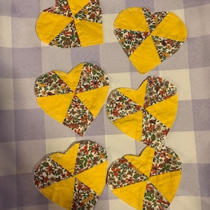 May include: Six heart-shaped fabric coasters featuring a patchwork design. Each coaster is constructed from alternating segments of solid bright yellow fabric and a floral print fabric containing small red, orange, and green flowers on a white background.