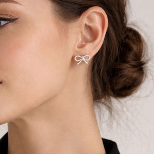 Puede incluir: Primer plano de una oreja adornada con pendientes de bot&oacute;n en forma de lazo. Cada pendiente est&aacute; adornado con peque&ntilde;os cristales brillantes, creando un efecto brillante. Los pendientes est&aacute;n colocados sobre la piel.