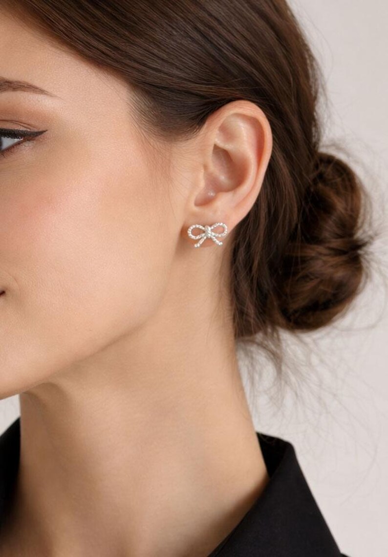 Puede incluir: Primer plano de una oreja adornada con pendientes de bot&oacute;n en forma de lazo. Cada pendiente est&aacute; adornado con peque&ntilde;os cristales brillantes, creando un efecto brillante. Los pendientes est&aacute;n colocados sobre la piel.