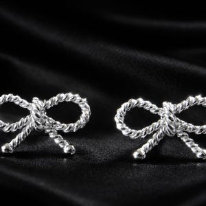 Puede incluir: Un par de pendientes de bot&oacute;n plateados con forma de lazo. Los pendientes est&aacute;n hechos de un material retorcido similar a una cuerda y est&aacute;n colocados sobre un fondo de sat&eacute;n negro. Los lazos son sim&eacute;tricos y detallados.