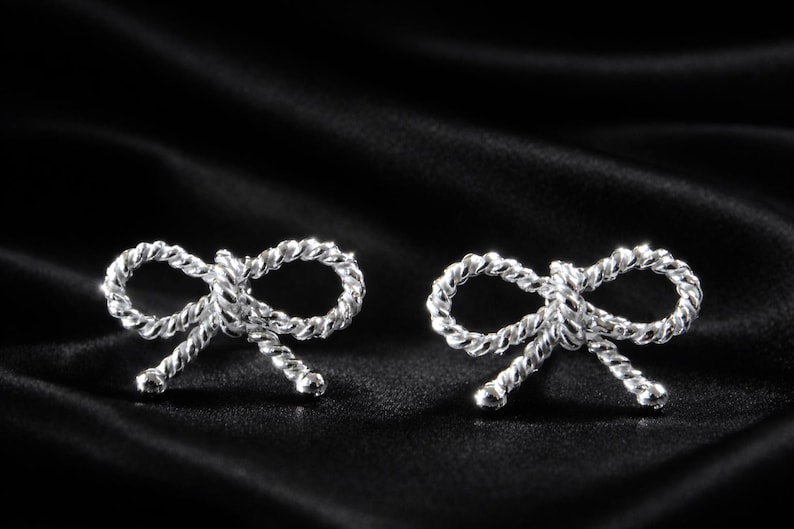 Puede incluir: Un par de pendientes de bot&oacute;n plateados con forma de lazo. Los pendientes est&aacute;n hechos de un material retorcido similar a una cuerda y est&aacute;n colocados sobre un fondo de sat&eacute;n negro. Los lazos son sim&eacute;tricos y detallados.