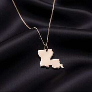 May include: A gold-colored necklace featuring a pendant shaped like the state of Louisiana. The pendant hangs from a delicate chain, set against a black satin background. The necklace is a simple, elegant piece of jewelry.