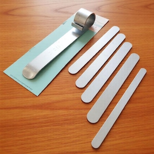 May include: A set of silver-colored metal tools, including a long, flat piece with a curved end and a clip, along with several thin, rectangular pieces. The tools are arranged on a light blue paper against a wooden surface.