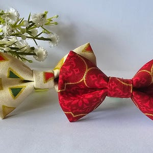 Pode incluir: Duas gravatas borboleta. Uma é creme com triângulos verdes, vermelhos e dourados. A outra é vermelha com um padrão geométrico dourado. Flores brancas ao fundo.