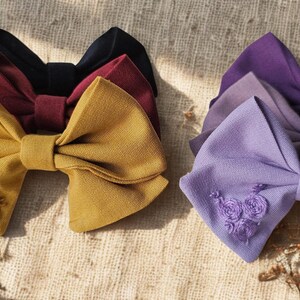 Pode incluir: Uma coleção de laços de cabelo de tecido em várias cores, incluindo amarelo mostarda, bordô, preto e tons de roxo. O laço amarelo apresenta detalhes florais bordados. Acessórios de cabelo.