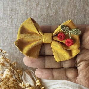 Pode incluir: Uma fita de tecido amarelo mostarda com um design de cereja. A fita tem duas cerejas vermelhas e folhas verdes. A fita é segurada na mão de uma pessoa.