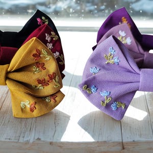 Pode incluir: Uma coleção de laços de cabelo de tecido em várias cores, incluindo amarelo mostarda, bordô, preto e tons de roxo. Cada laço é adornado com delicados bordados florais, adicionando um toque de elegância. Os laços estão dispostos em uma superfície de madeira branca.