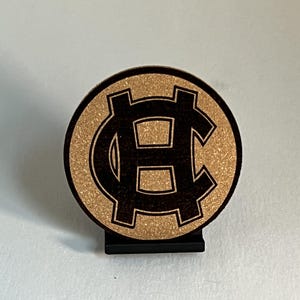 May include: A round, wooden coaster with a dark brown monogram design, set against a textured, light brown background. The coaster is displayed on a small, black stand, suitable for a desk or table.