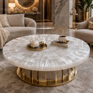 May include: A round, white coffee table with a radial pattern on the surface. The table has a gold-colored metal base. Decorative items, including a crystal cluster and a small tray, are on the table's surface. The table is in a living room setting.
