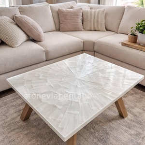 May include: Square white coffee table with a radial pattern, set on a light wood base. The table is in front of a beige corner sofa. The surface has a distinctive, textured design. The table measures approximately 90cm x 90cm.