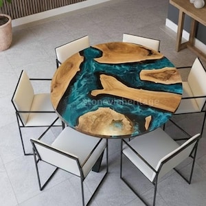Op de afbeelding: Een ronde eettafel met een houten blad en een turquoise hars rivierontwerp, omringd door zes witte stoelen met zwarte metalen frames. De diameter van de tafel is ongeveer 122 cm.