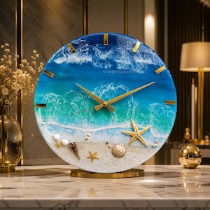 Könnte beinhalten: Eine runde Wanduhr mit einer Harz-Ozeanszene, die blaue und türkisfarbene Wellen zeigt, die an einen Sandstrand branden. Das Zifferblatt enthält goldfarbene Stundenmarkierungen, goldene Zeiger sowie eingebettete Muscheln und Seesterne.