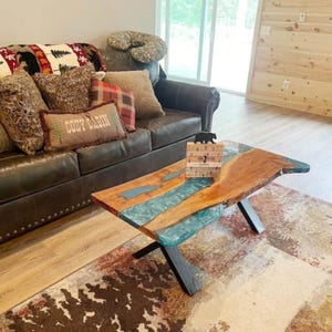 May include: A rustic coffee table with a live edge design. The table features a wooden surface with a teal epoxy resin river. The base is made of black metal. A small wooden block with a bear silhouette and text is on the table.