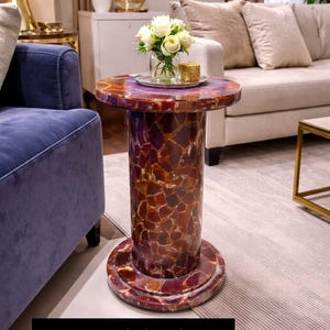 May include: A decorative side table with a mosaic-style design. The table features a round top with a purple surface and a cylindrical base. The base and sides are a mosaic of red, brown, and purple stones. The text "stoneviaheritage" is at the bottom.