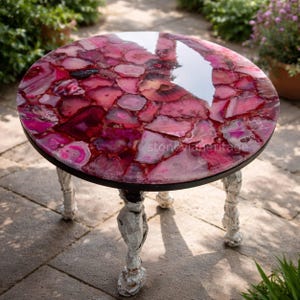 May include: A round side table with a pink agate stone top. The table has a black metal frame and ornate white legs. The agate top features shades of pink and red. The table is a decorative piece for a living room or patio.