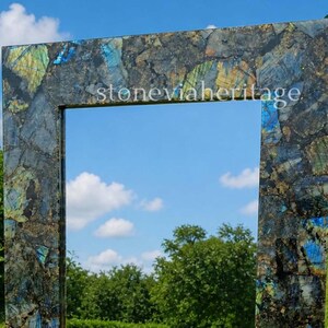 Puede incluir: Espejo rectangular con marco de mosaico de piedras azules, verdes y marrones. El texto "stoneviaheritage" es visible en la parte superior del marco. El espejo refleja un cielo azul con nubes blancas y &aacute;rboles verdes.