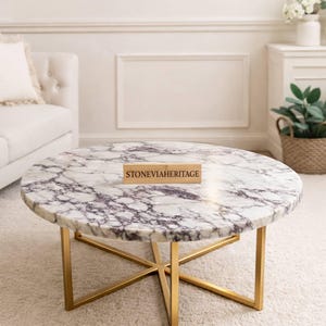 Op de afbeelding: Een ronde salontafel met een wit marmeren blad met grijze adering. De tafel heeft een goudkleurig metalen onderstel. De tekst "STONEVIAHERITAGE" staat op een houten bord op de tafel.