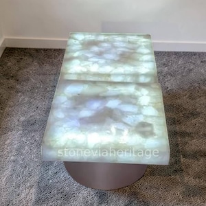 May include: A rectangular coffee table with a glowing, translucent top made of white and light blue stones. The table is supported by a round, metallic base. The text "stoneviaheritage" is visible on the table's edge.