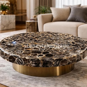 Puede incluir: Una mesa de centro redonda con una tapa de piedra oscura estampada y una base dorada. La tapa de piedra presenta un diseño tipo mosaico con tonos marrones, negros y crema. La mesa es un punto focal en un salón moderno.