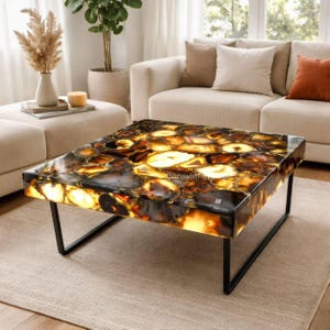 May include: A square coffee table with a polished, agate stone top in shades of brown, amber, and black. The table has a black metal frame. The room has a neutral color palette with a beige sofa and rug.