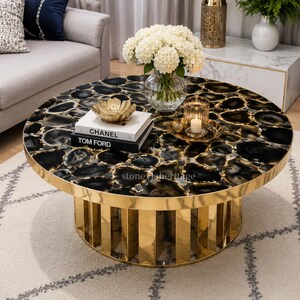 May include: A round coffee table with a black and gray agate stone top and a gold-colored metal base. The table has a gold rim and a cylindrical base with vertical gold bars. Decorative items include books, a gold bowl, and a candle.