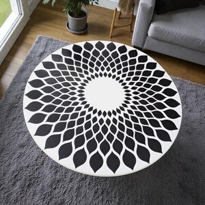 May include: A round white coffee table with a black floral pattern. The table's design features a central white circle surrounded by a repeating black leaf-like motif. The table is placed on a gray rug.