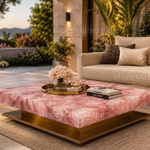 Puede incluir: Una mesa de centro cuadrada de cuarzo rosa con base dorada. Una bandeja dorada con flores y libros encima. La mesa está en un entorno al aire libre con un sofá y plantas.