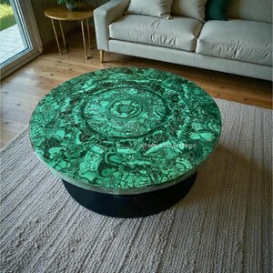 Puede incluir: Una mesa de centro redonda con una llamativa tapa de malaquita verde. La superficie presenta un diseño arremolinado único en tonos verdes y negros. La base es un cilindro negro macizo. La mesa descansa sobre una alfombra de color neutro.