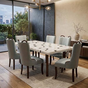 May include: A rectangular dining table with a white stone top featuring dark, irregular spots, supported by dark wooden legs. Six upholstered dining chairs in a light blue-grey fabric with dark wooden frames surround the table. Text reads stoneviaheritage.