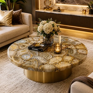 Puede incluir: Una mesa de centro redonda con una superficie de piedra pulida que presenta un mosaico de colores neutros y detalles dorados. La mesa tiene una base de metal dorado. Un libro con la palabra "CHANEL" está encima.