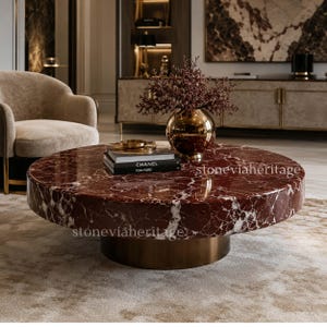 May include: A circular coffee table with a red marble top and a brass base. The marble features white veining. The table is in a living room setting with a beige armchair and a gold vase.