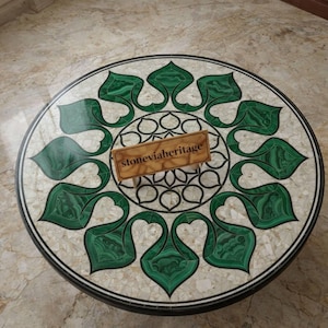 May include: Round marble table with a decorative inlaid design featuring green leaf-like shapes and a central floral pattern. The table has a black border and a light-colored background. The text "stoneviaheritage" is on a wooden sign.