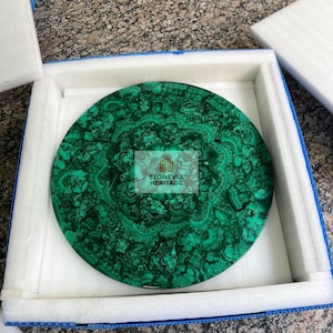 May include: A round, polished malachite stone with intricate green patterns. The stone is displayed in a white foam-lined box with a blue border. The text "STONEVIA HERITAGE" is visible on the stone.