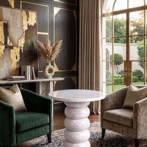 Op de afbeelding: Een witte marmeren bijzettafel met een gestapelde ringvoet staat tussen een donkergroene fluwelen fauteuil en een taupekleurige fluwelen fauteuil. De stoelen hebben gouden sierkussens en staan op een blauw en goud patroon vloerkleed in een kamer.