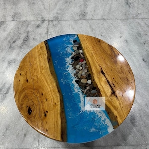 May include: Round wooden coffee table with a blue epoxy resin river design. The river is filled with small stones and shells, creating a coastal theme. The table has a natural wood grain finish and a glossy top.