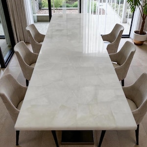 May include: A large, rectangular dining table with a white, patterned surface. The table is surrounded by six beige upholstered chairs. The table's dimensions appear to be approximately 100 inches long, suitable for a dining room setting.