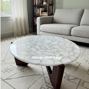 May include: A round coffee table featuring a white quartz stone top with a mosaic-like pattern of irregular pieces and a smooth, polished finish. The table is supported by three dark brown wooden legs with a V-shaped design. Text reads stoneviaheritage.
