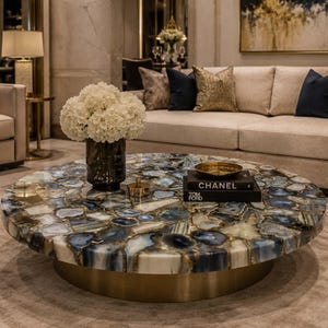 May include: A round coffee table with a mosaic top featuring blue, white, and gray stones. The table has a gold-colored base and holds a vase of white flowers, a gold bowl, and books.