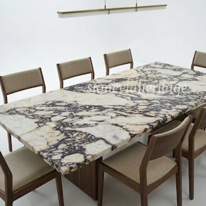 Puede incluir: Una mesa de comedor rectangular con una tapa de mármol con un patrón blanco y morado oscuro. La mesa está rodeada de ocho sillas de madera con asientos y respaldos tapizados en beige. Una moderna lámpara cuelga encima.