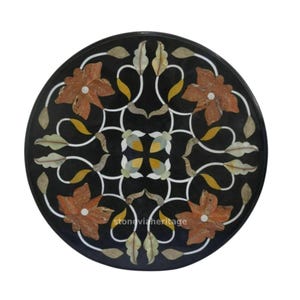 Op de afbeelding: Ronde zwarte marmeren tafelblad met een ingelegd bloemmotief. Het ontwerp heeft oranje, gele en witte accenten. De tekst "stoneviaheritage" is zichtbaar op de tafel.