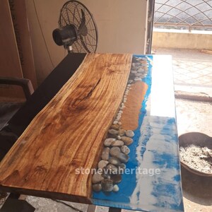 May include: A handcrafted wooden table with a unique design. The table features a natural wood grain surface on one side and a resin design on the other, with a beach-themed aesthetic. The resin incorporates blue, white, and brown elements, with small stones along the edge.