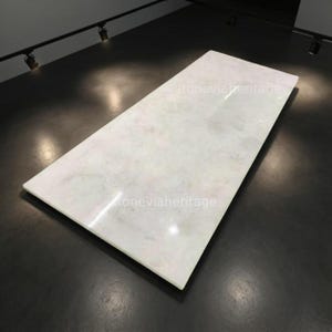 Può includere: Una lastra rettangolare di marmo bianco con venature sottili. La superficie lucida riflette la luce, creando un aspetto lucido. Il testo "stoneviaheritage" è visibile sulla superficie. La lastra è posta su uno sfondo scuro e contrastante.