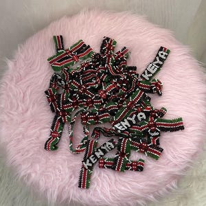 May include: A pile of beaded accessories in red, black, green, and white, resting on a pink, fluffy surface. Some pieces feature the word "KENYA" in white beads. The accessories appear to be handmade.