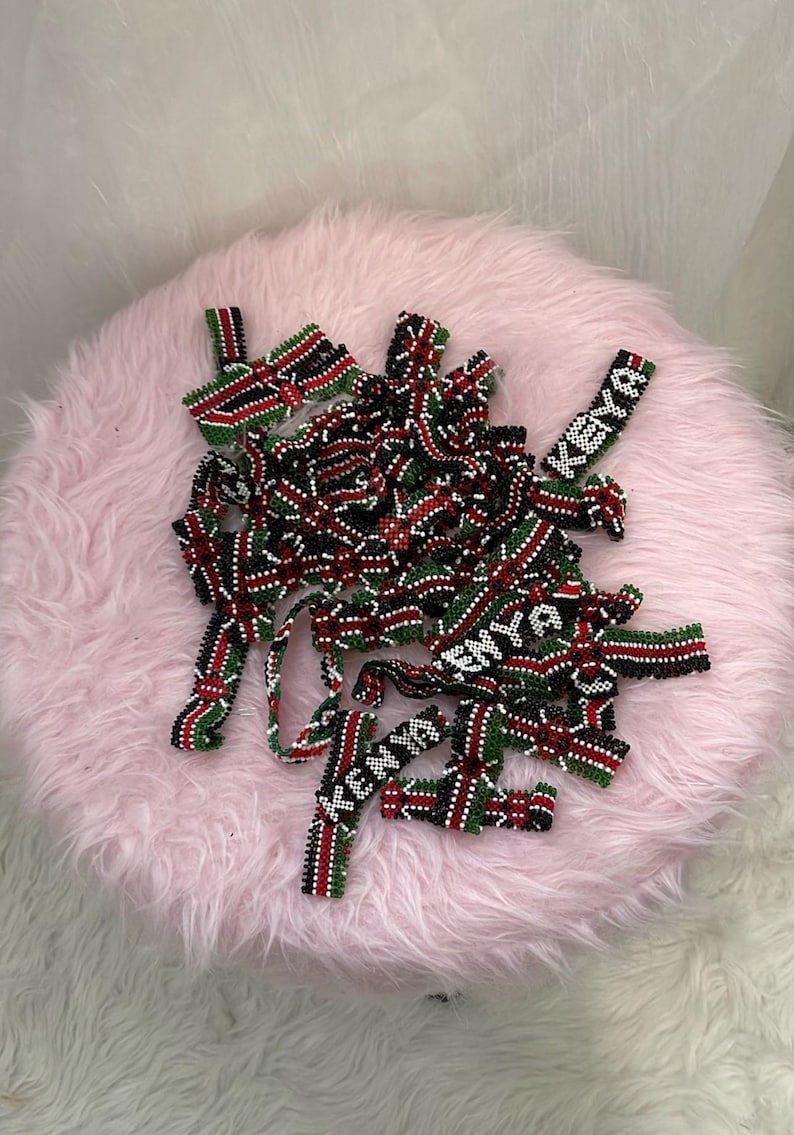 Puede incluir: Un conjunto de accesorios de cuentas rojas, negras, verdes y blancas, colocados sobre una superficie rosa y esponjosa. Algunas piezas tienen la palabra "KENYA" en cuentas blancas. Los accesorios parecen hechos a mano.